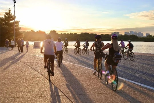 A group of people ride bicycles and walk along a paved riverside path at sunset, with trees and a city skyline in the background.