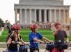 Three people wearing helmets with bicycles in front of the Lincoln Memorial, with the man pointing and crowds visible on the steps behind them.