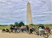 A group of people with bicycles and a bike trailer pose in front of the Washington Monument on a cloudy day.