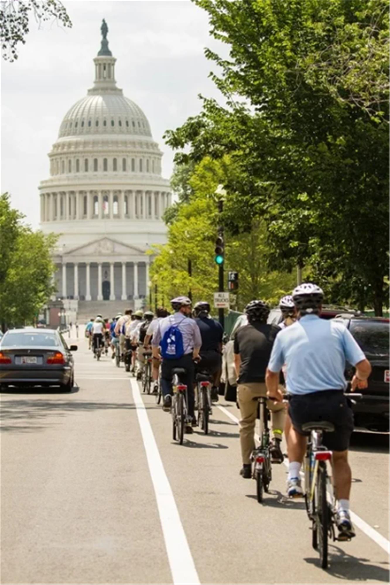 A group of cyclists ride along a bike lane toward the U.S. Capitol building on a sunny day, passing cars parked on the street.