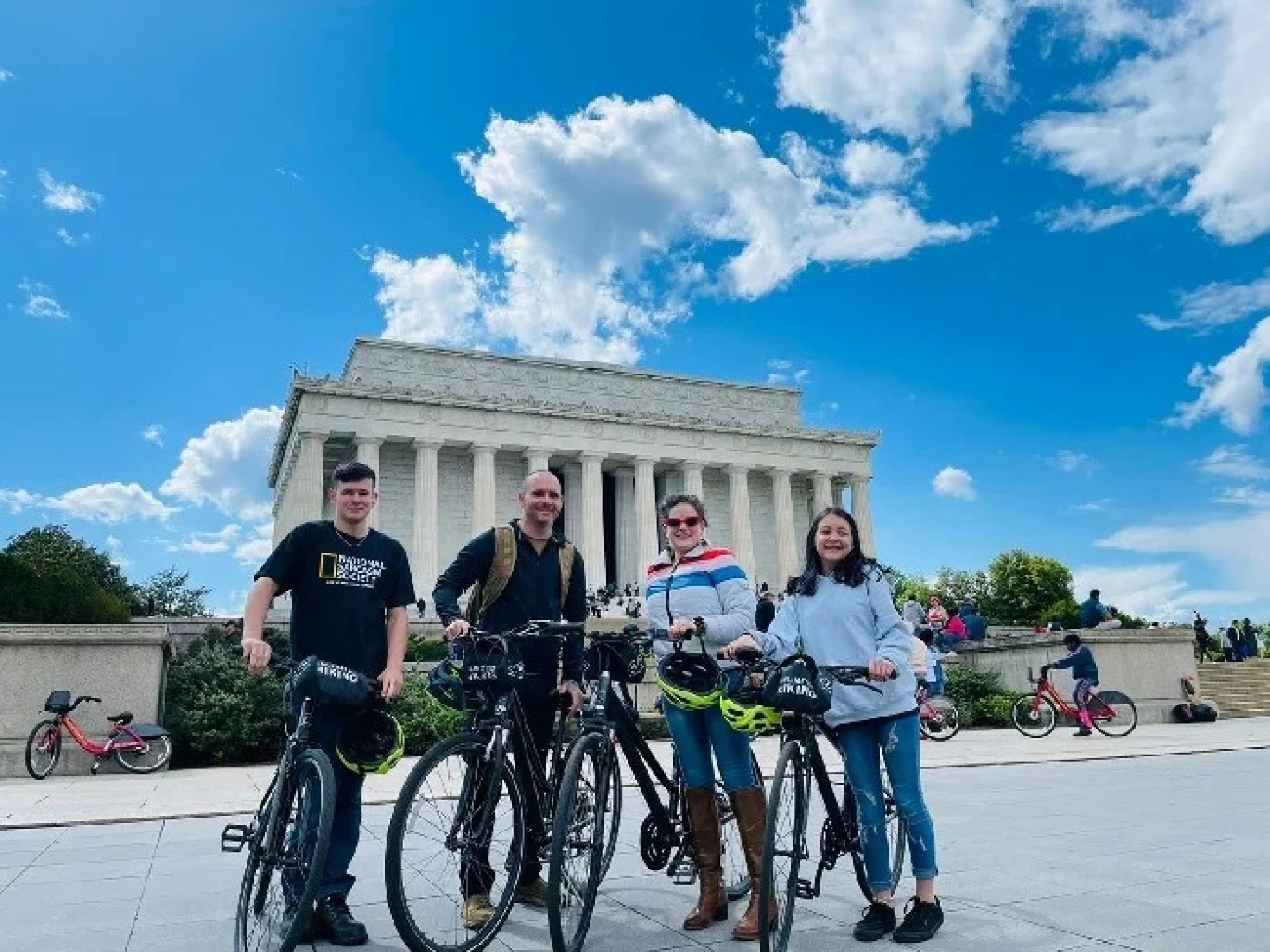Four people with bicycles pose in front of the Lincoln Memorial on a sunny day with a blue sky and scattered clouds.