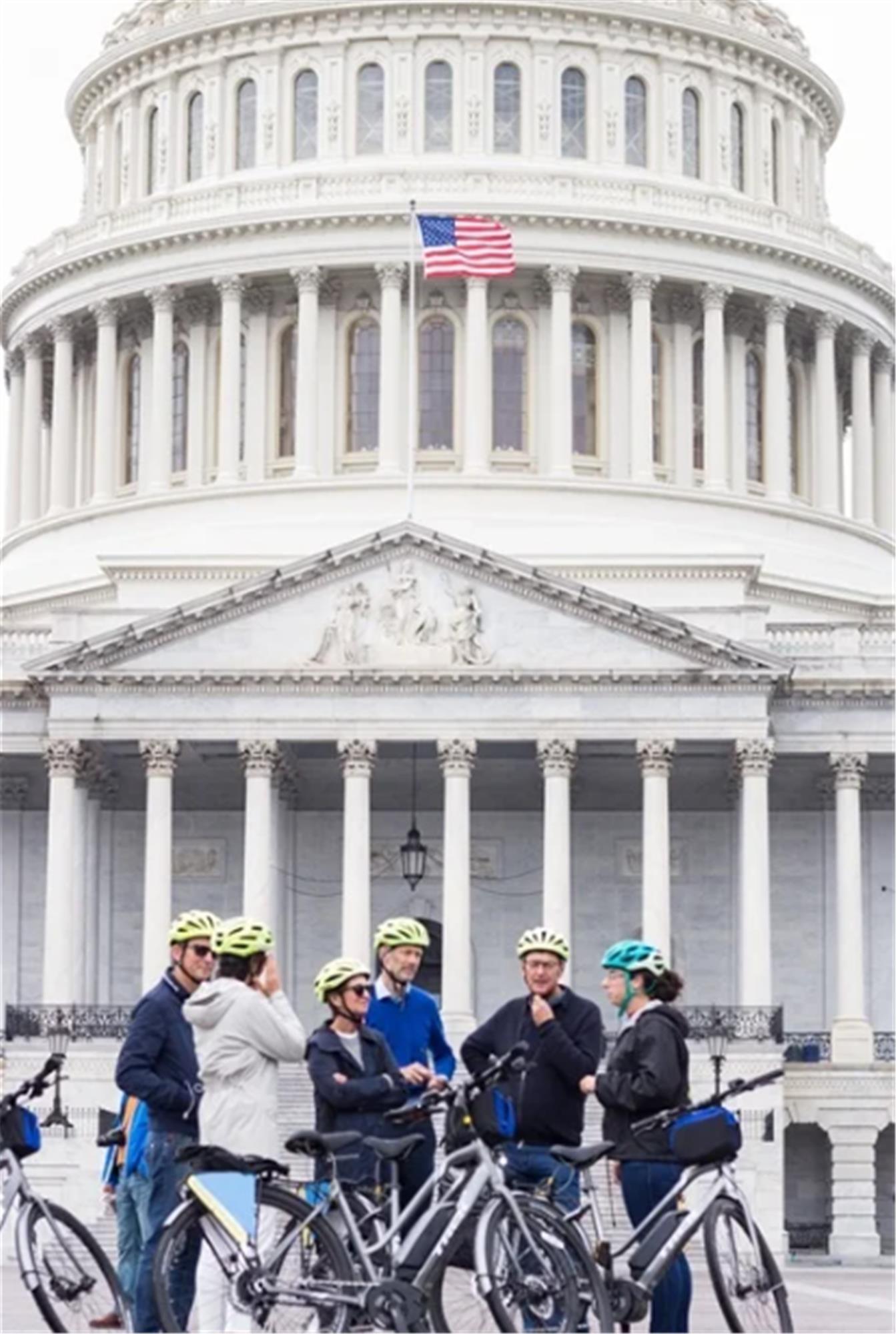 A group of people with bicycles and helmets gather in front of the U.S. Capitol building, with the American flag visible above the entrance.
