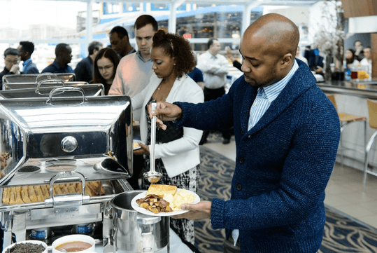 People serving food from a buffet with chafing dishes at an indoor event.