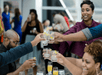 People clink glasses in a toast at a social gathering, with drinks and lively conversation evident.