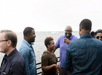 Group of people standing and talking near a waterfront, with sailboats visible in the background. One person is holding a drink.