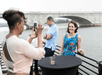 Woman in a floral dress poses for a photo taken by another woman with a smartphone. A man stands nearby. They're on a riverside deck with a bridge in the background.