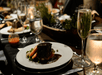 A plate of braised beef with vegetables is served on a restaurant table. The table features multiple glasses of water and champagne, along with decorative greenery and a candle.