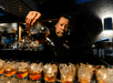 Bartender pouring liquor into a line of glasses filled with ice at a dimly lit bar.