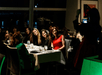 A group of people seated at a dining table pose for a photo. A person standing nearby takes the picture, while others smile and face the camera.