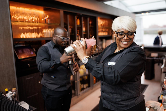 Bartenders in black attire prepare cocktails at a bar; one shakes a pink drink in a metal shaker, while the other fills a glass with a beverage. Glassware and bottles are displayed behind them.