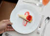 A hand serving a slice of cheesecake topped with red cherries on a white plate, next to cutlery and a glass with a pink drink.