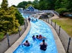People float on blue inner tubes along a winding lazy river at a water park, surrounded by trees, rocks, and a blue building in the background.