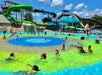 Children and adults swim and play in a shallow pool at a water park with large water slides in the background under a partly cloudy sky.