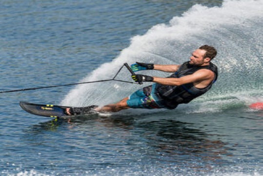 A person water skiing on a lake, wearing a life jacket and holding a tow rope, with water spray and a buoy visible in the background.