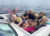 Nine children in life jackets are sitting in a boat on a lake, smiling and posing for the camera.