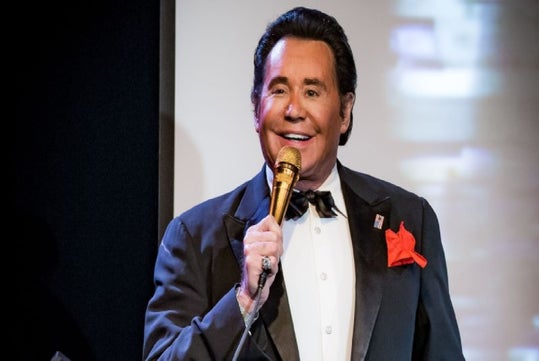 Man in a tuxedo and red pocket square singing into a gold microphone on stage.