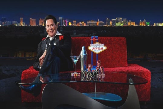 Wayne Newton sitting on a red sofa with Las Vegas as the background.