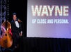 Wayne Newton sitting on a red chair with a stand up bass next to him and a projector screen in the background.