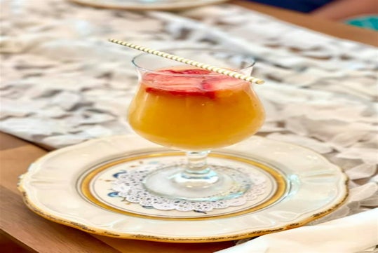 A glass of orange-colored beverage with ice and sliced strawberries, served on a decorative plate with a lace doily and a yellow striped straw.