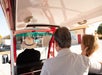 Three people ride in a small, open vehicle; the driver wears a wide-brimmed hat, and two passengers sit in the back, facing forward toward buildings outside.