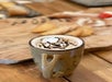 A ceramic mug filled with hot chocolate topped with whipped cream and chocolate drizzle sits on a wooden table, with pastries and powdered sugar in the background.