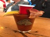 A Bloody Mary cocktail garnished with lemon, lime, a strip of bacon, and a mini red cup with a blue straw, served in a glass featuring a goat illustration.