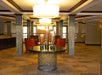 Hotel lobby with a round table in the center, featuring tall vases with red flowers. Surrounded by red chairs and illuminated by large overhead lights.