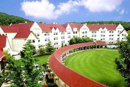 A large building with a red roof surrounded by green trees and a grassy lawn under a partly cloudy sky. A curved colonnade leads to the entrance.