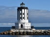 A black and white lighthouse stands on a rocky base surrounded by water under a partly cloudy sky.