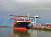 A large container ship is docked at a port under towering cranes, with a colorful rainbow arcing across the cloudy sky in the background.
