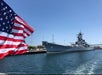 US Navy battleship number 61 docked at a harbor, with an American flag in the foreground and clear blue sky overhead.