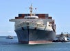 A large container ship docked at a port is assisted by a tugboat, with another smaller vessel visible in the background under a clear blue sky.