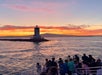 A group of people on a boat views a lighthouse on a rocky outcrop at sunset, with orange and pink clouds over calm water and distant hills.