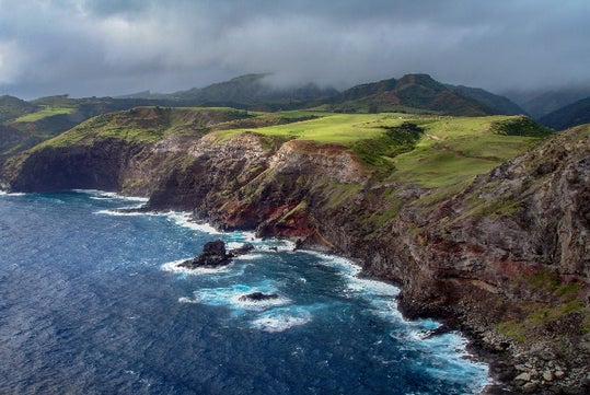 Rocky coastline with steep cliffs and green grassy hills overlooking a deep blue ocean under a cloudy sky.