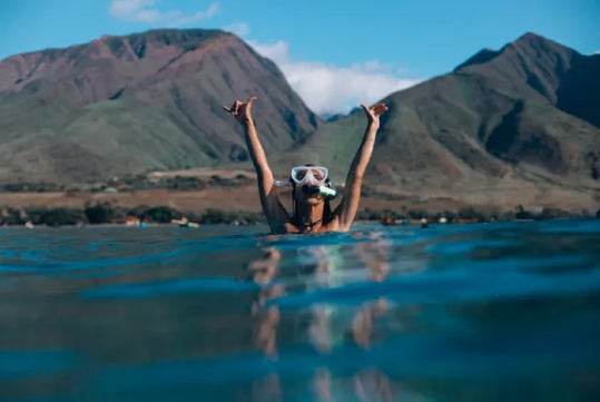 A person wearing snorkel gear raises their arms above the water with mountains in the background under a clear blue sky.