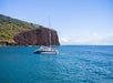 A white catamaran floats on clear blue water near a rocky, green cliff under a sunny sky.