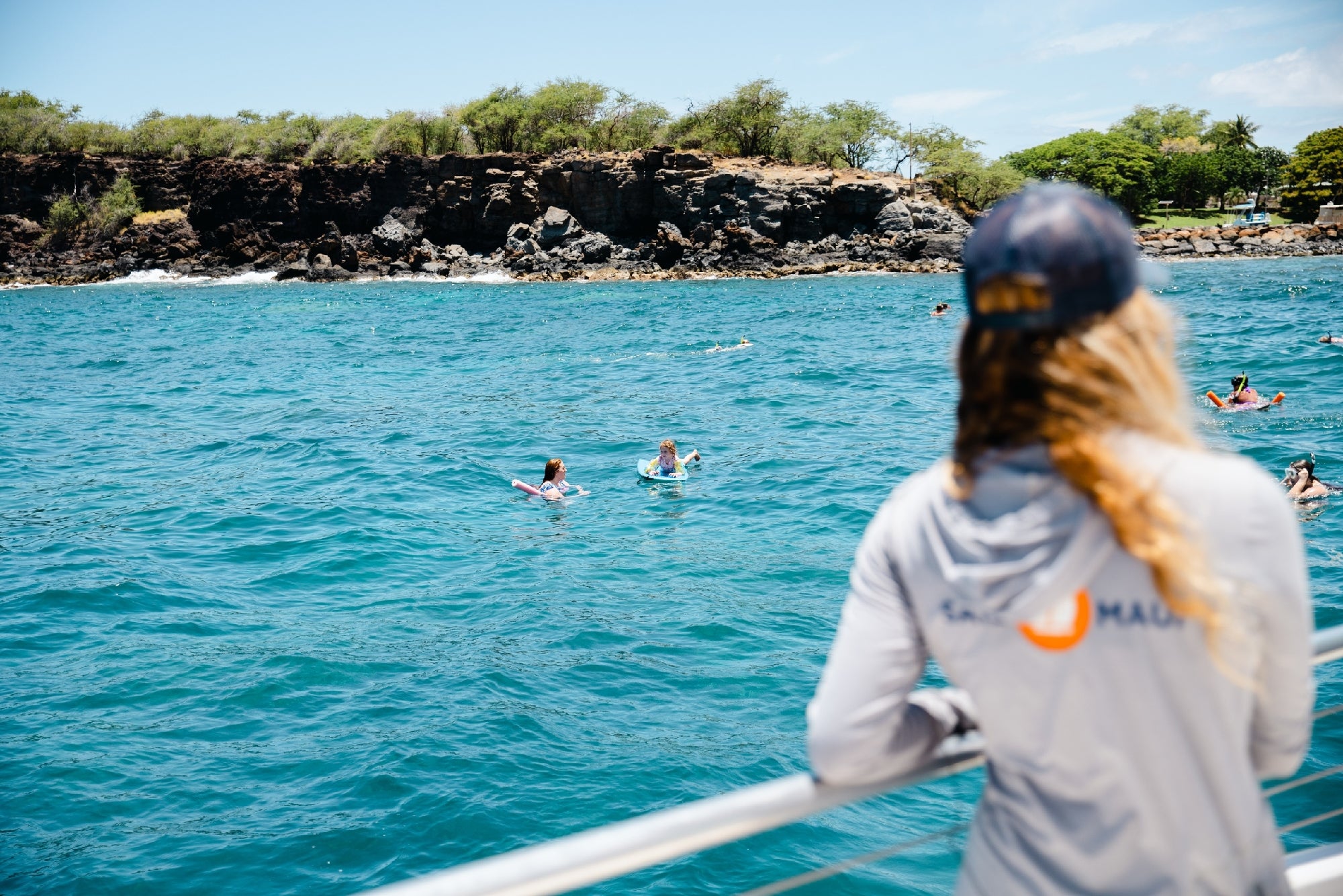A person stands on a boat watching swimmers in blue ocean water near a rocky shoreline with trees under a partly cloudy sky.