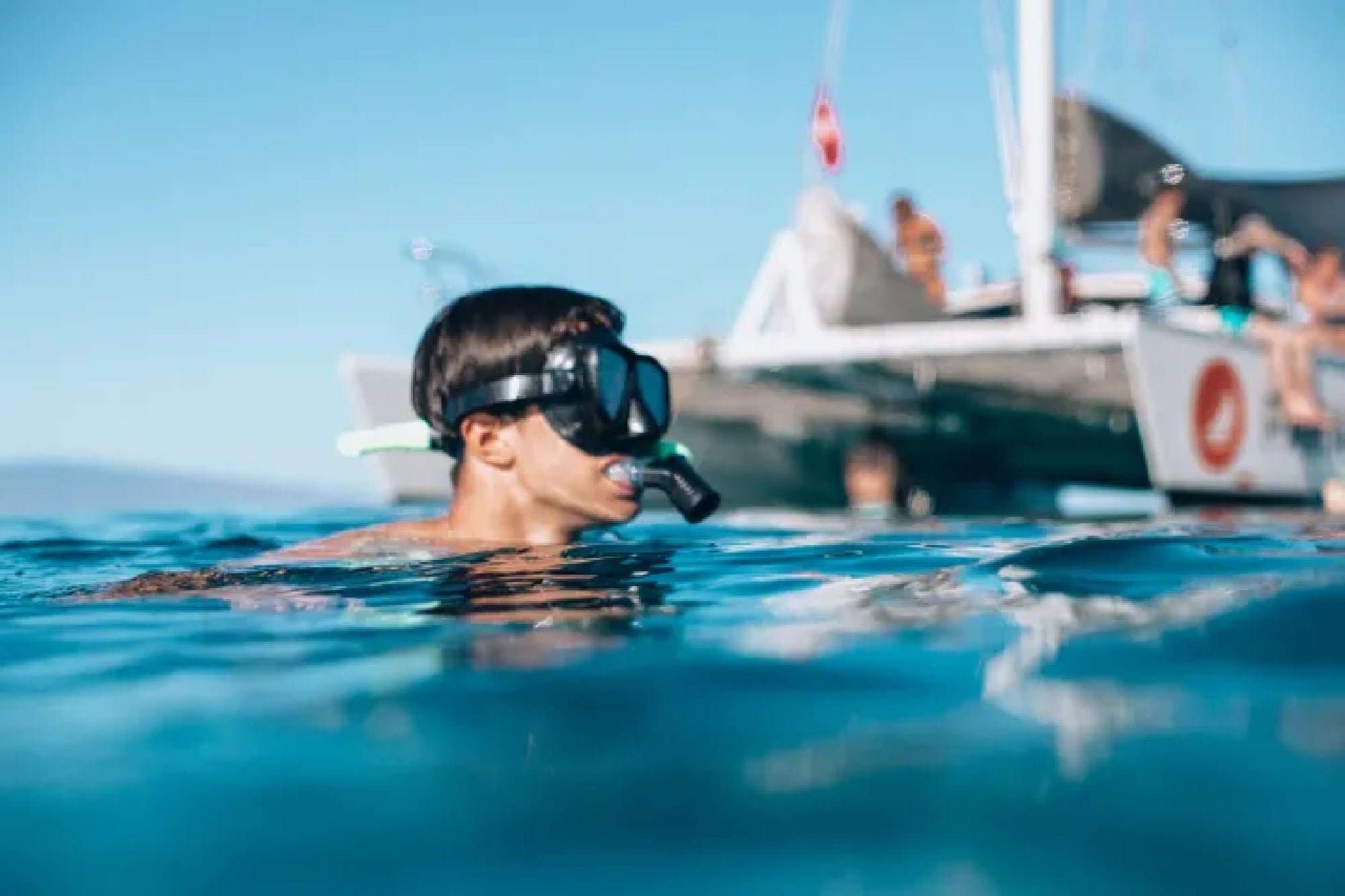 Person wearing snorkel mask swims near a boat with people on board, in clear blue water under a sunny sky.