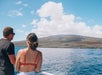 Two people stand on a boat looking toward a mountainous coastline under a partly cloudy sky with blue ocean water in the foreground.