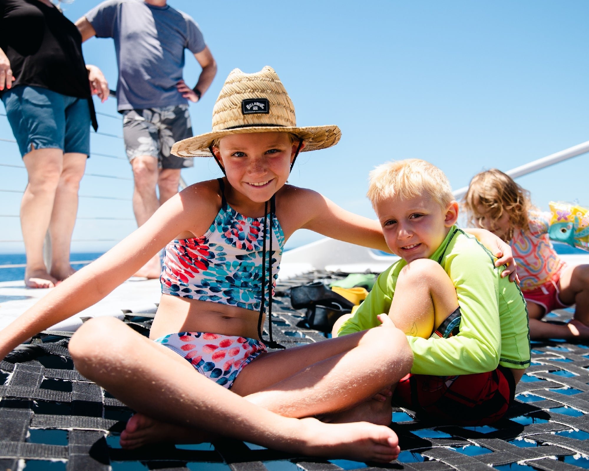 Two children sit together on a netted deck in swimwear, with adults and another child in the background under a clear blue sky.