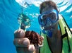 A snorkeler wearing a yellow life vest holds a sea urchin underwater, with clear blue water in the background.