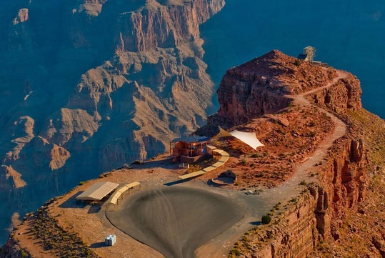 Aerial view of a cliffside tourist area with a building, walkways, and a curved viewing platform, overlooking rugged red rock formations under a clear blue sky.