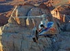 A silver helicopter flies above a rocky desert landscape with large sandstone formations and rugged terrain in the background.