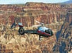 A silver helicopter flies over the Grand Canyon, with layered rock formations visible in the background.