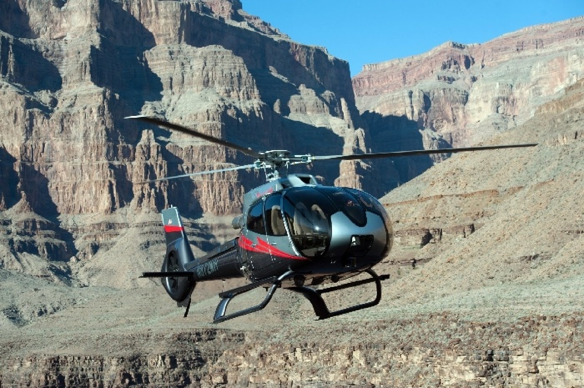A helicopter flies above rocky terrain with steep cliffs and canyon walls in the background under a clear blue sky.