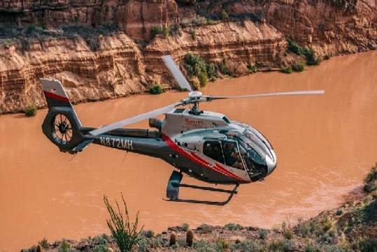 A helicopter with red and gray markings flies above a muddy river in a rocky canyon landscape.