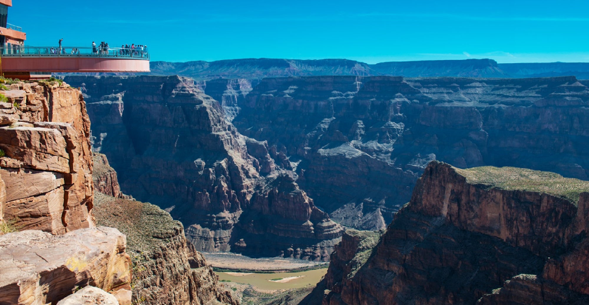 A glass skywalk extends over the edge of the Grand Canyon with visitors standing on it, overlooking the deep canyon and river below under a clear blue sky.