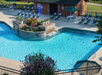 Outdoor swimming pool with lounge chairs, tables with umbrellas, a small waterfall feature, and a nearby building. A sign and potted plants are visible around the pool area.