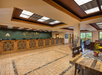 Hotel lobby with a wooden reception desk, tiled floor, decorative ceiling, and seating area with plants. A sign on the wall reads "Westgate Branson Woods.