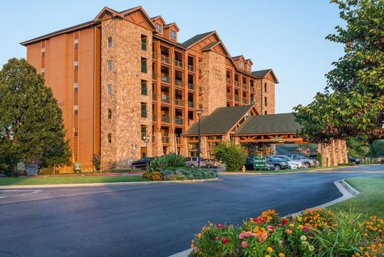 A multi-story hotel with stone and wood facade, surrounded by trees and colorful flowers in the foreground. Several cars are parked near the entrance.